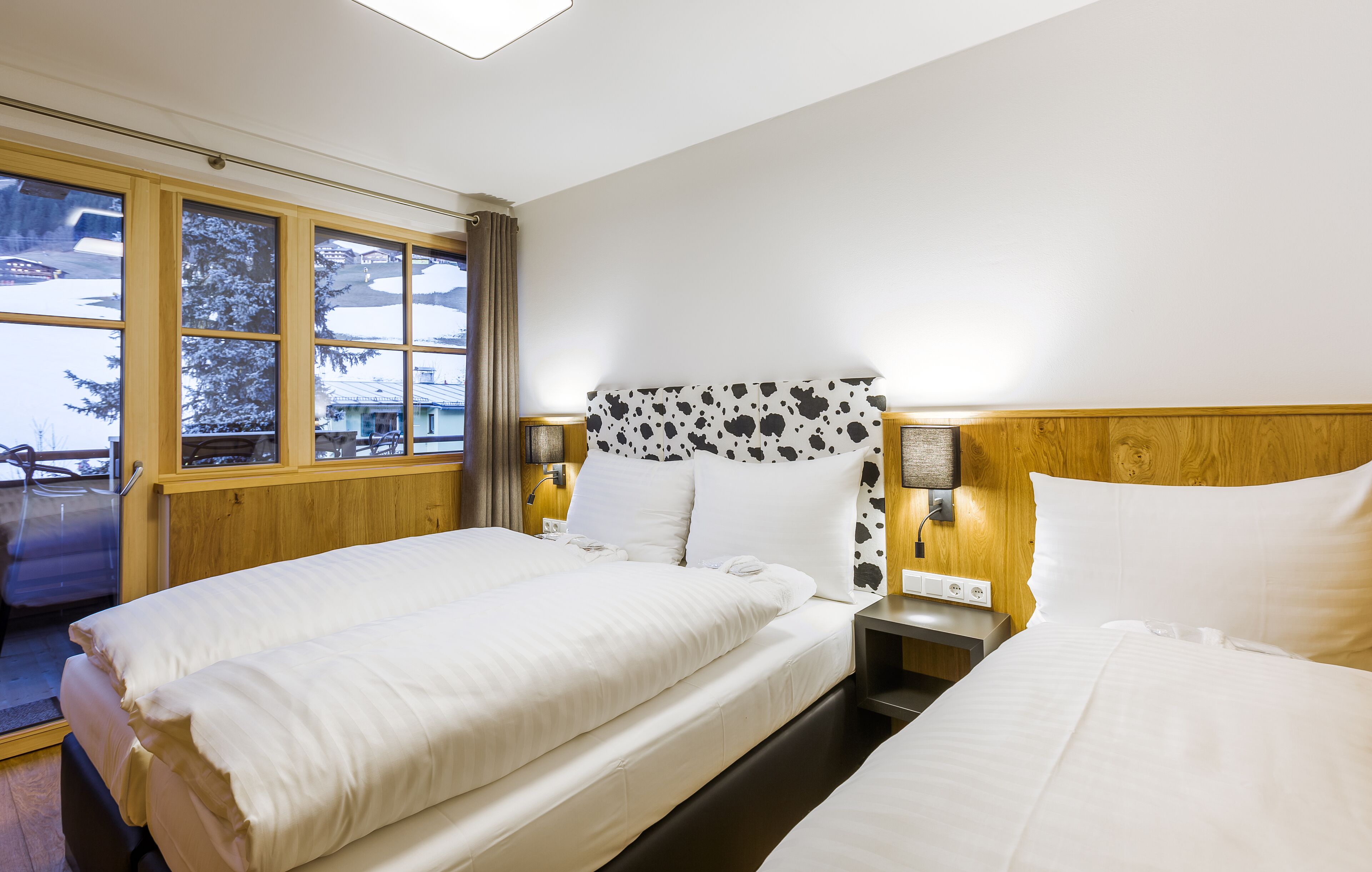 A hotel room with two beds, a wooden headboard, a nightstand, and a window with a view of snow-covered trees.