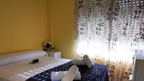 A double bed with a dark blue bedspread and white pillows, a small bedside table with a vase of flowers, and a window with floral curtains. - Charming Apartment with Pool Access, Sea Views and Terrace in Playa de Aro (Platja d'Aro)