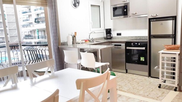 A modern kitchen with stainless steel appliances, a dining area with white furniture, and a view of a balcony with a pool. - Charming Apartment with Pool Access, Sea Views and Terrace in Playa de Aro (Platja d'Aro)