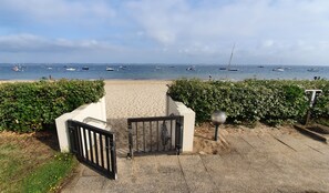 Uma entrada para a praia com um portão, um sebedeiro e vista clara de barcos na água.