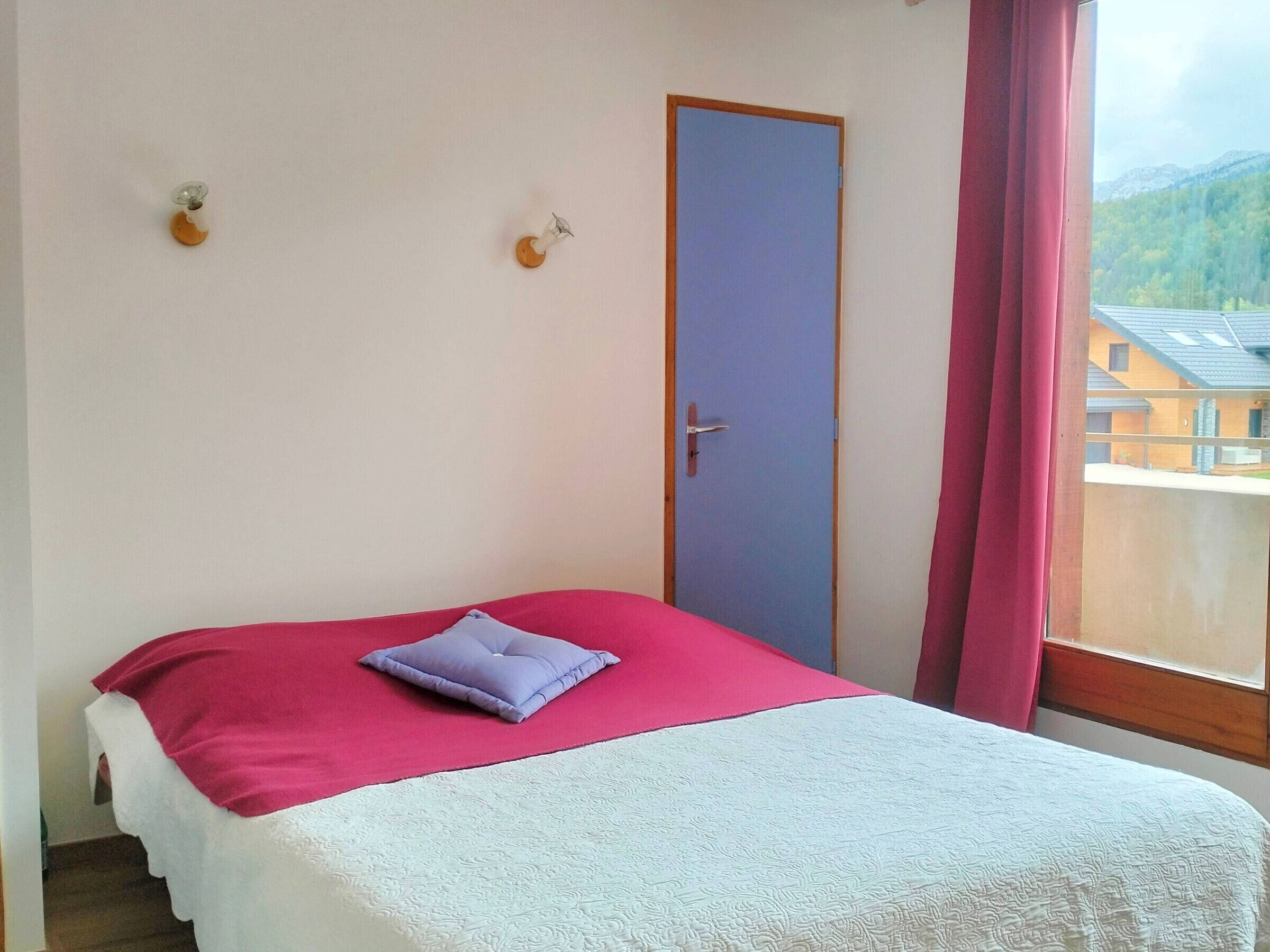A bedroom with a bed, a door, and a window with a view of buildings and mountains.