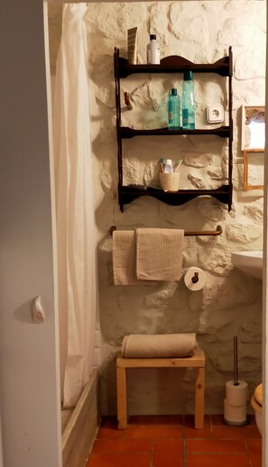 Ein Badezimmer mit Steinwand, Holzregal mit Toilettenartikeln, Handtuchhalter, einem kleinen Holzschemel und einem Toilettenpapierhalter.