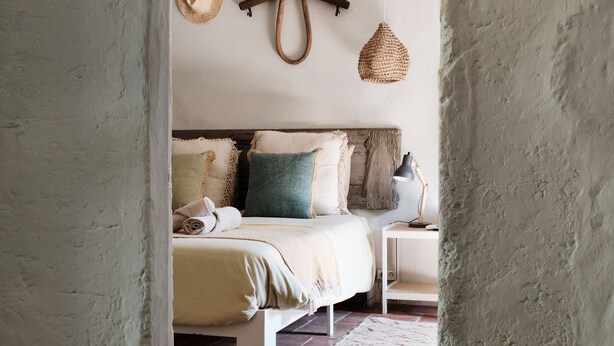 A rustic bedroom with a bed, pillows, a hanging light fixture, and a woven basket on the wall.
