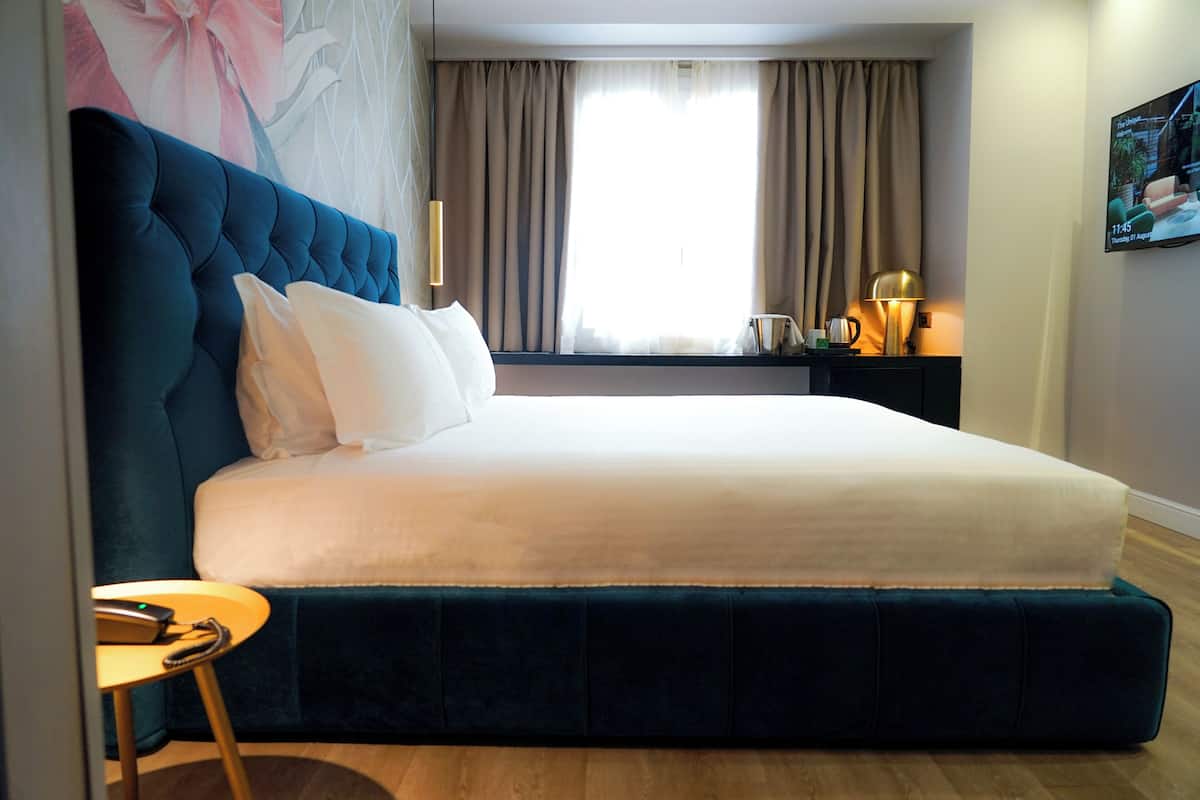 A modern hotel room with a large bed, a bedside table with a lamp, and a view of the outdoors through the window.
