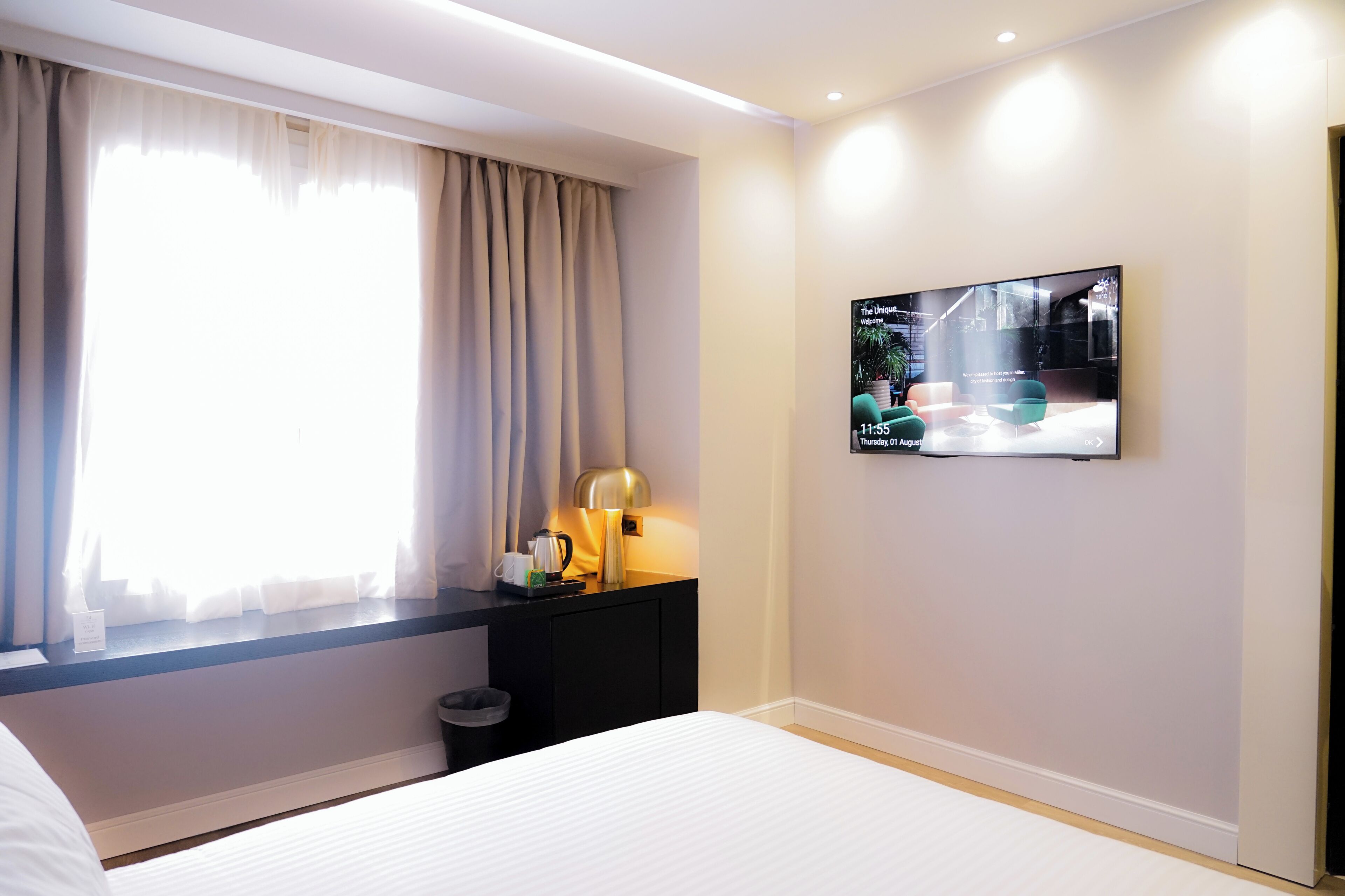 Ein Hotelzimmer mit Bett, Schreibtisch mit Lampe, Fernseher an der Wand und einem Fenster mit Vorhängen.