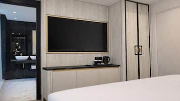 Quarto de hotel moderno com uma cama grande, televisão de tela plana na parede, mesa de cabeceira pequena com cafeteira e banheiro privativo com pia e espelho.