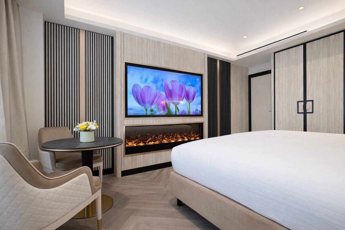 A modern hotel room with a large bed, a flat-screen TV mounted on the wall, a small round table with a vase of flowers, and a built-in fireplace.