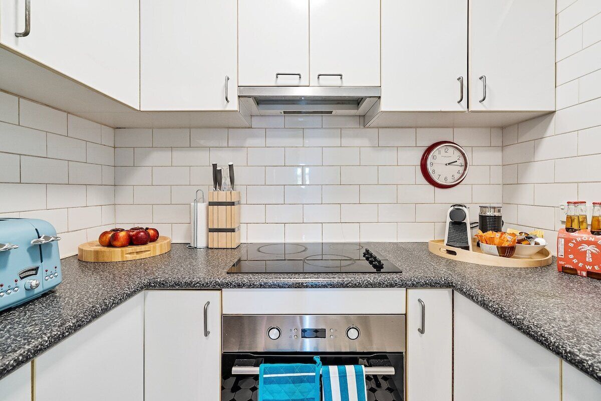 A modern kitchen with white cabinets, a stainless steel range hood, a built-in oven, a clock, and a coffee machine.