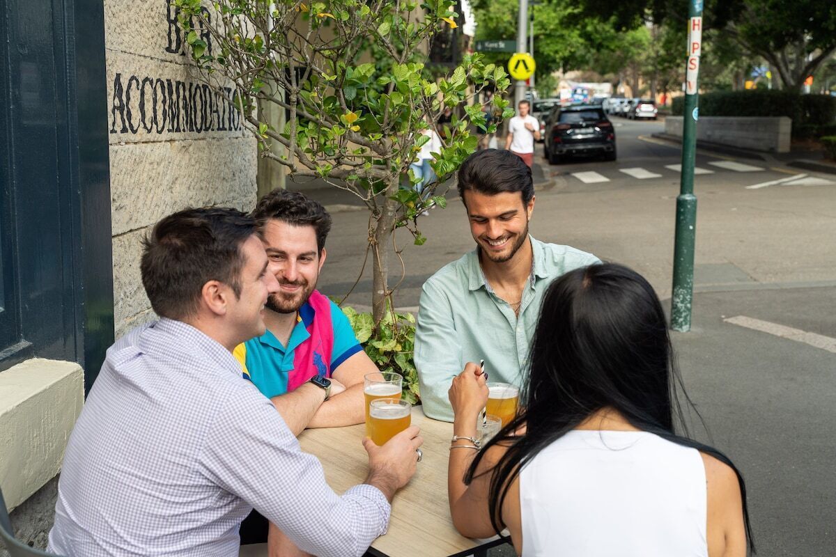 A group of people sitting at an outdoor table, holding glasses of beer, with a building labeled "ACCOMMODATION" in the background.