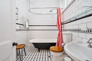 Un baño con una bañera blanca, un lavamanos blanco y un inodoro. Hay un taburete de madera y una toalla blanca y con rayas rojas colgada en un perchero.