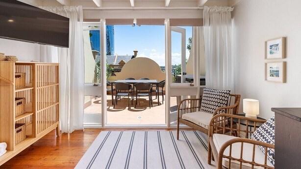 Una habitación con estantería de madera, sofá de mimbre, mesa de comedor con sillas y vistas a un patio exterior a través de un ventanal.
