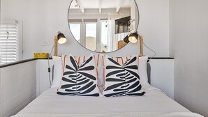 Una cama con ropa blanca y dos almohadas de estampado blanco y negro, un espejo redondo encima de la cama, y dos lámparas de noche.