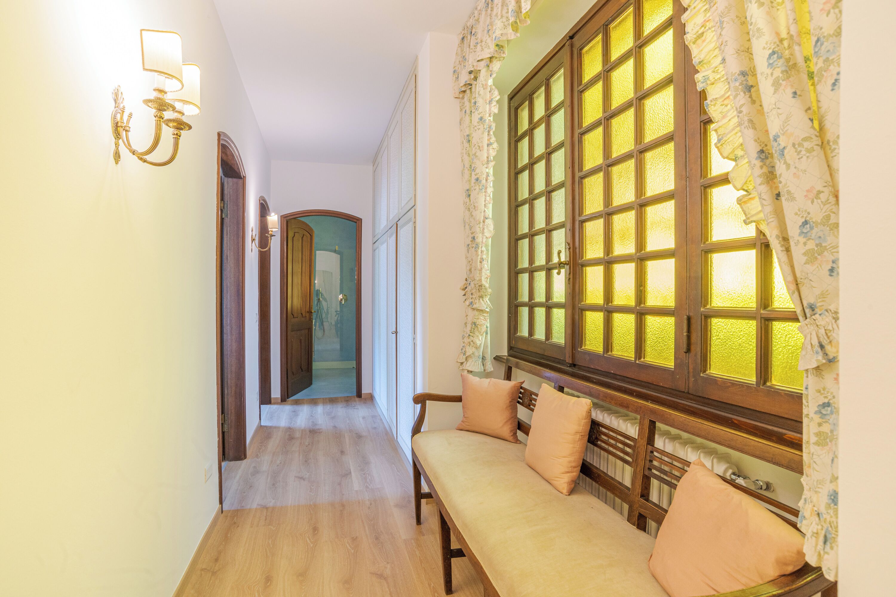 A hallway with a wooden bench, a wall-mounted light fixture, and a window with floral curtains.