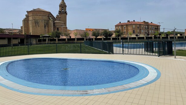 Una piscina con borde de baldosas, rodeada por una valla, y un edificio histórico con una torre de campanas al fondo.