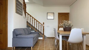 Una sala de estar con un sofá gris, una mesa de comedor de madera con sillas blancas, una escalera con barandilla de madera y una puerta con marco de madera.