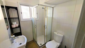 Ett badrum med en dusch som är avskärmad med glasväggar, en toalett, ett handfat och en hylla med toalettpapper.
