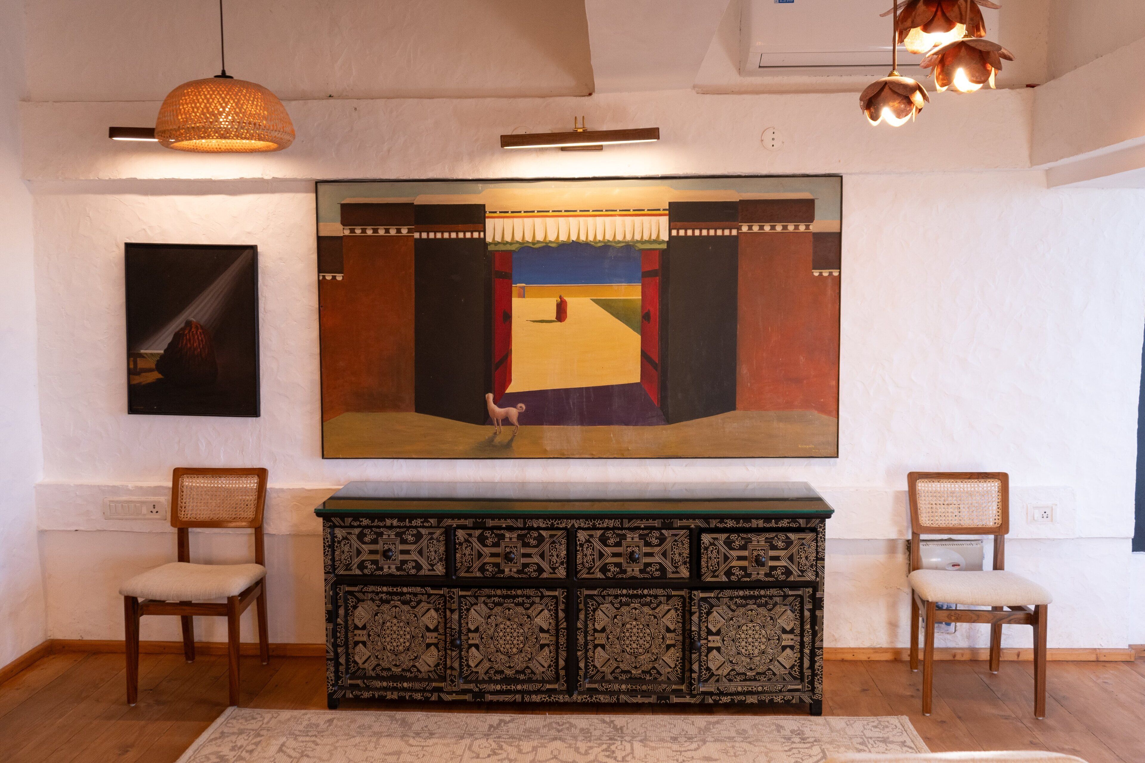 Une pièce avec un grand tableau, deux chaises en bois, un meuble orné de motifs et des lampes suspendues.