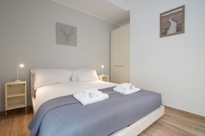 A neatly made bed with a grey quilt, white pillows, and folded towels on a wooden floor.