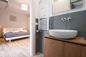 A modern bedroom with a bed, bedside tables, a large mirror, and a bathroom with a sink and towel rack.