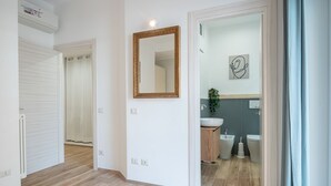 Ein Flur mit Holzboden, einer weißen Tür, einem Spiegel sowie einem Badezimmer mit Waschbecken, Bidet und Toilette.
