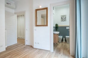 A hallway with a wooden floor, a white door, a mirror, and a bathroom with a sink, bidet, and toilet.