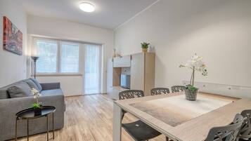Ett modernt vardagsrum med en grå soffa, ett matbord med stolar, ett litet sidobord med en växt och ett köksområde med skåp och en tv.