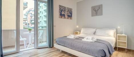 Un dormitorio con cama, mesita de noche, lámpara fijada a la pared y puerta corrediza de vidrio que da a un balcón con mobiliario exterior.