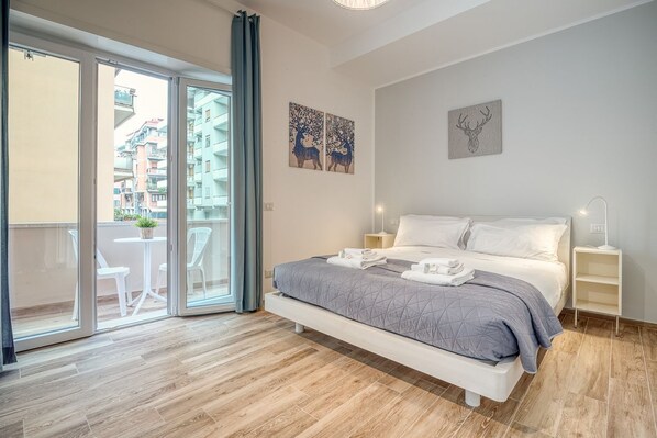 A bedroom with a bed, a nightstand, a wall-mounted lamp, and a sliding glass door leading to a balcony with outdoor furniture.