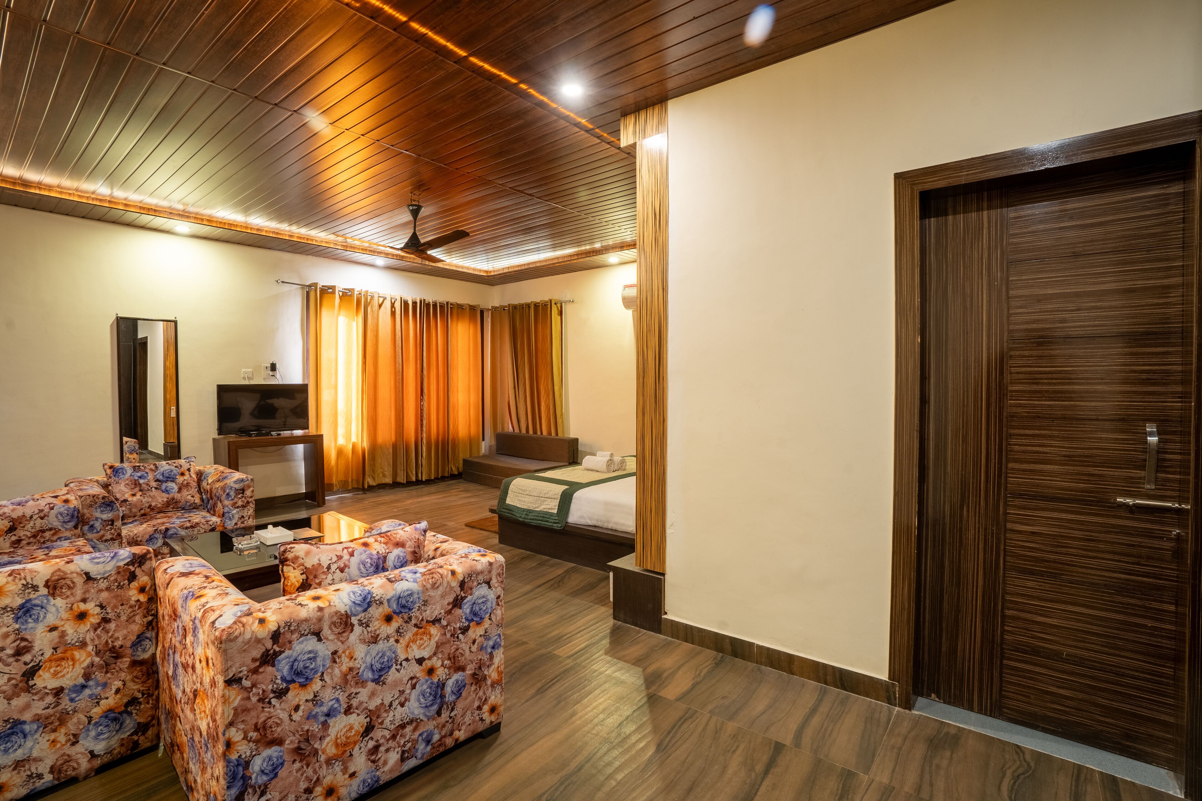 A hotel room with a wooden ceiling, a bed, a sofa, a TV, and a wooden door.