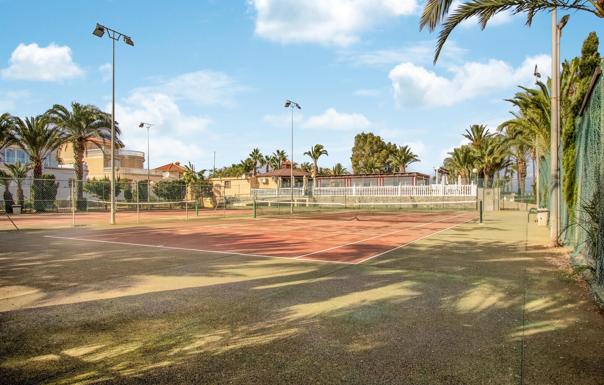 Una cancha de tenis exterior con red, rodeada de palmeras y edificios.