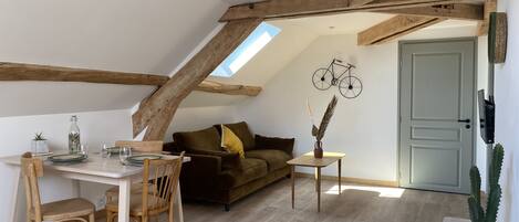 A cozy loft-style room with exposed wooden beams, a dining area with a table and chairs, a sofa, and a small side table with a vase.