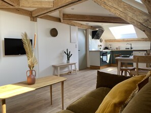 A cozy loft-style living space with exposed wooden beams, a dining area, a kitchen with a modern design, and a large skylight. - Appartement de Charme au Pied du Grand Colombier (CULOZ)