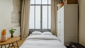 Un dormitorio con cama, una mesita de noche con una planta, un armario y una ventana con cortinas.