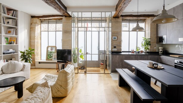 Una sala moderna con sofá, una mesa de centro y una estantería.  Se observa una zona de cocina con una mesa de comedor y bancos.