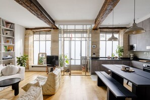 Uma sala de estar moderna com um sofá, uma mesa de centro e uma estante de livros. Há uma área de cozinha com uma mesa de jantar e bancos.