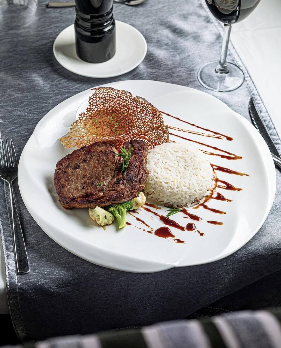 A plate with a steak, rice, and vegetables, garnished with sauce, on a dark table.