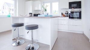 A modern kitchen with white cabinets, a central island, two bar stools, and stainless steel appliances. - Modernes Ferienhaus in Attraktiver Lage in Burhave. Strandnah und Zentral (Burhave)