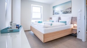 Un dormitorio moderno con una cama de madera, ropa de cama blanca, una mesita de noche con una lámpara y una ventana con vista a la playa.