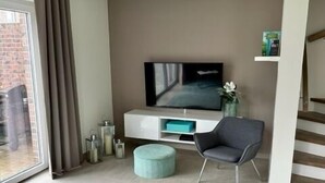 A modern living room with a flat-screen TV, a grey armchair, a teal ottoman, and a patterned rug. - Modernes Ferienhaus in Attraktiver Lage in Burhave. Strandnah und Zentral (Burhave)