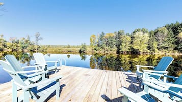 Une terrasse en bois avec quatre chaises Adirondack donnant sur un lac paisible, entouré d’arbres.
