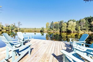 Eine Holzterrasse mit vier Adirondack-Stühlen, die auf einen ruhigen See mit umliegenden Bäumen ausgerichtet ist.