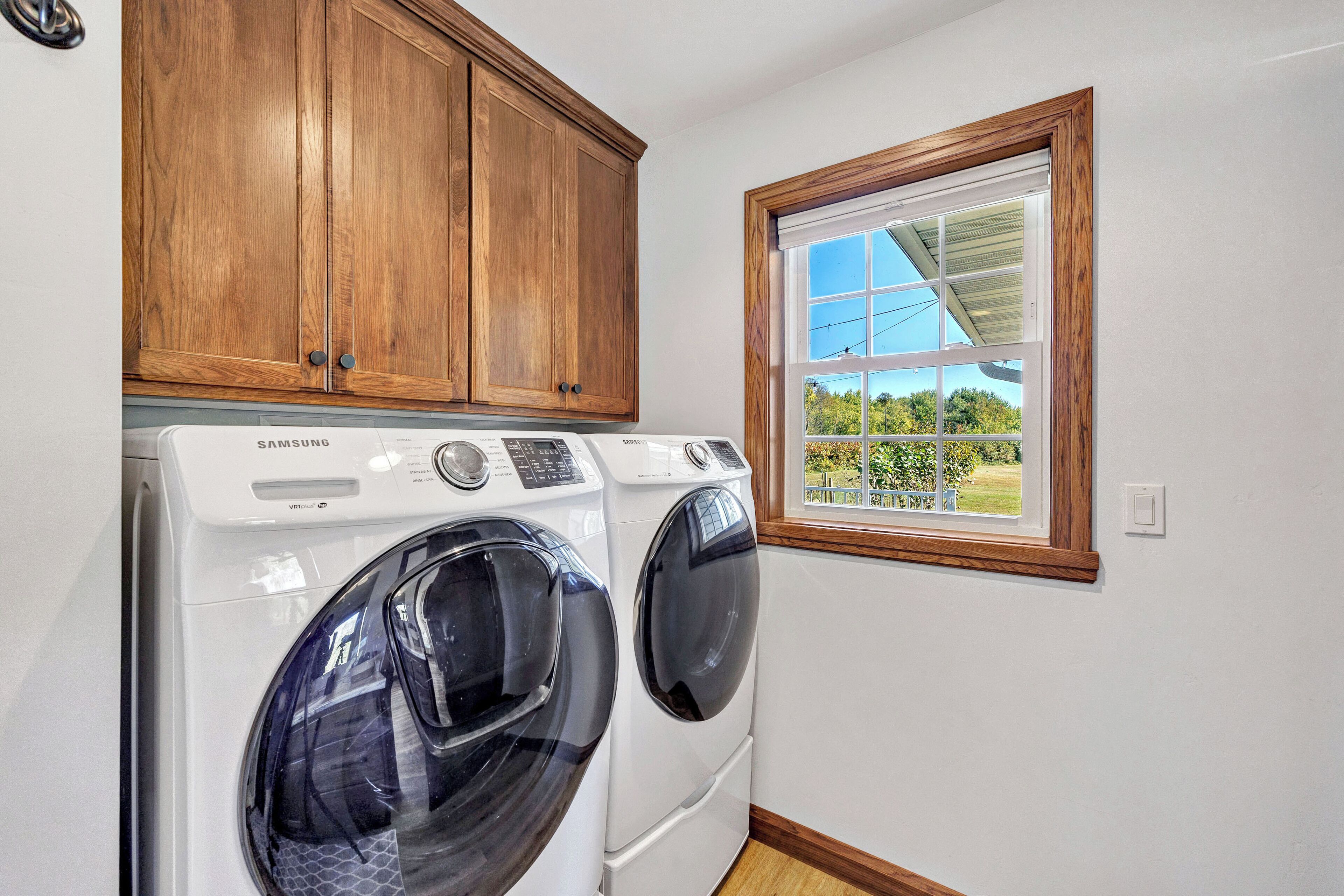A laundry room with two front-loading washing machines, wooden cabinets, and a window with a view of greenery.