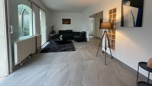 A modern living room with a black sofa, a small table with a plant, and a lamp on a tripod stand. There is a radiator on the left and a wall with a painting and a shelf with decorative items.