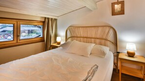 A bedroom with a wooden ceiling, a bed with white bedding, a window with curtains, and a bedside table with a lamp. - Charming Apartment for 6 in the Heart of Les Gets Village, Close to Slopes (Les Gets)