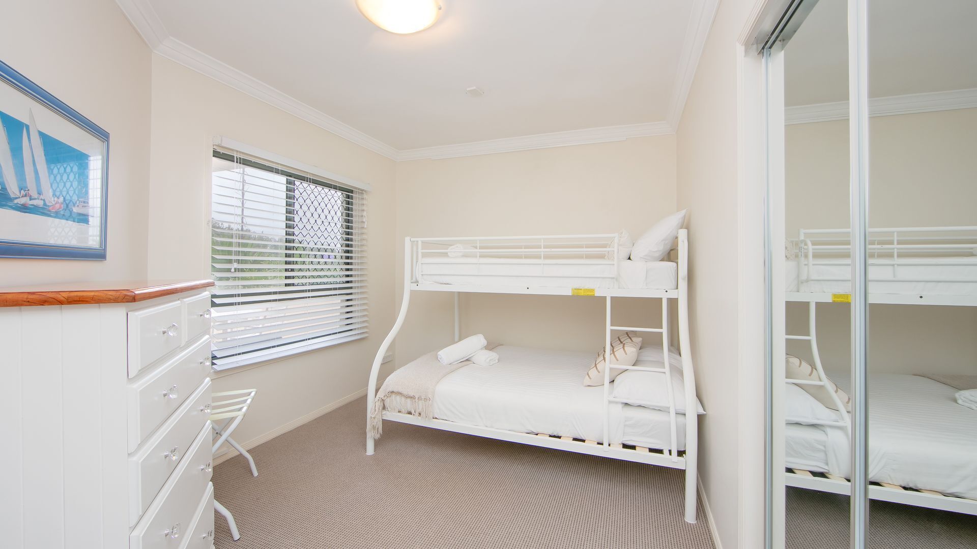 A bunk bed room with a desk and chair, a window with blinds, and a framed picture on the wall.