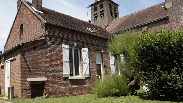 Un bâtiment en briques doté d’un clocher, d’un petit porche et d’un espace verdoyant agrémenté d’arbustes.