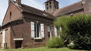 Un edificio de ladrillo con un campanario, un pequeño porche y un área ajardinada con arbustos.