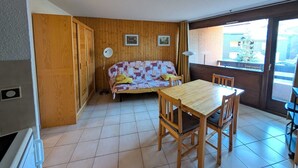 Een compacte kamer met een houten vloer, een houten tafel met vier stoelen, een bank met een sprei en een raam met uitzicht op gebouwen en een boom.