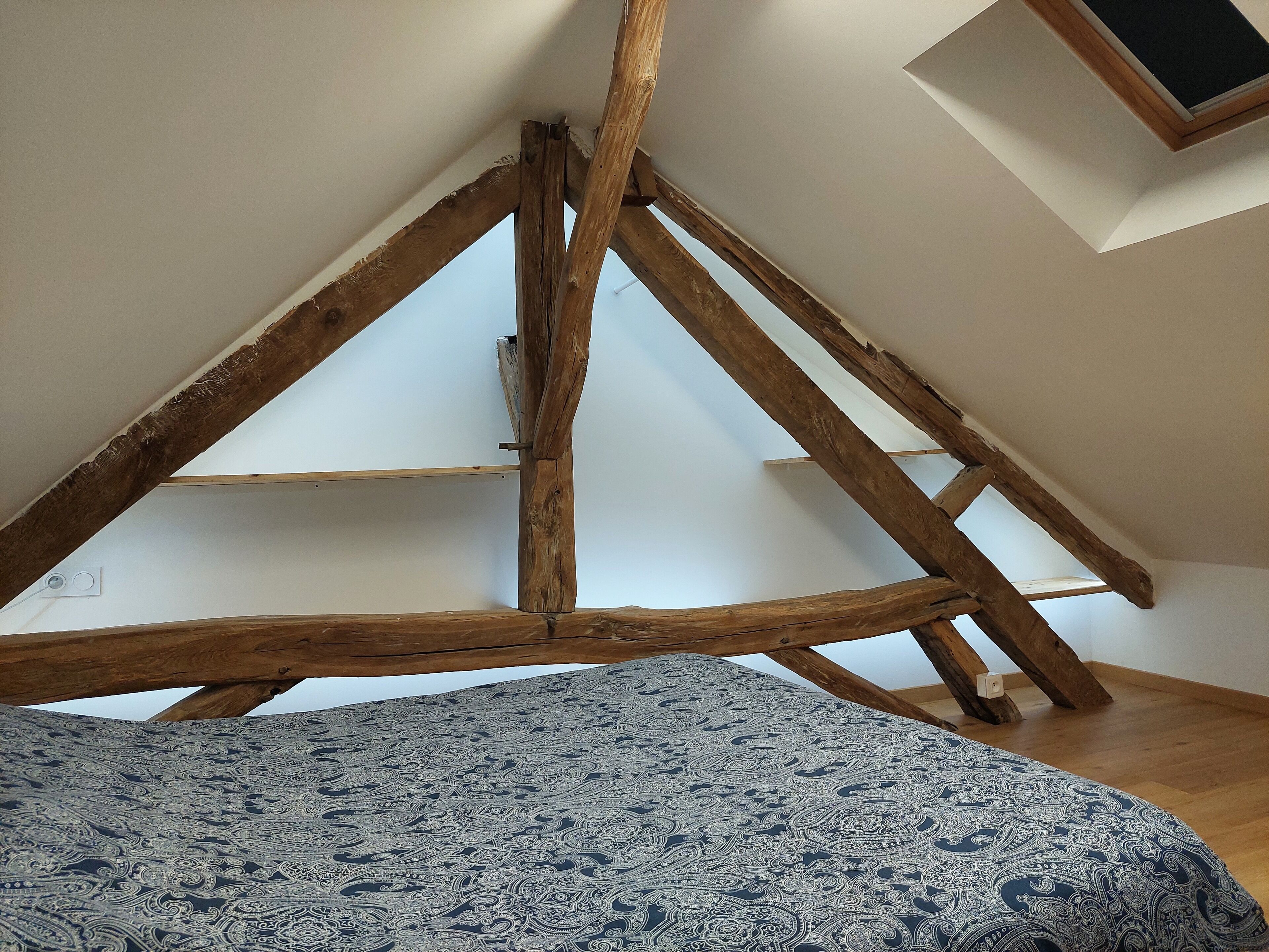 A room with a sloped ceiling supported by wooden beams, a patterned bedspread, and a small window.