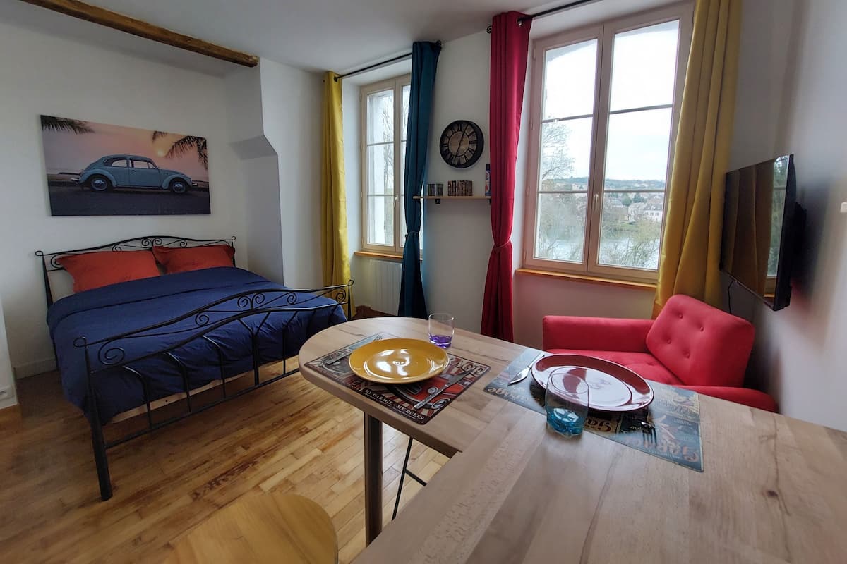 A bedroom with a bed, a table with a plate and a glass, a red chair, a window with curtains, and a clock on the wall.